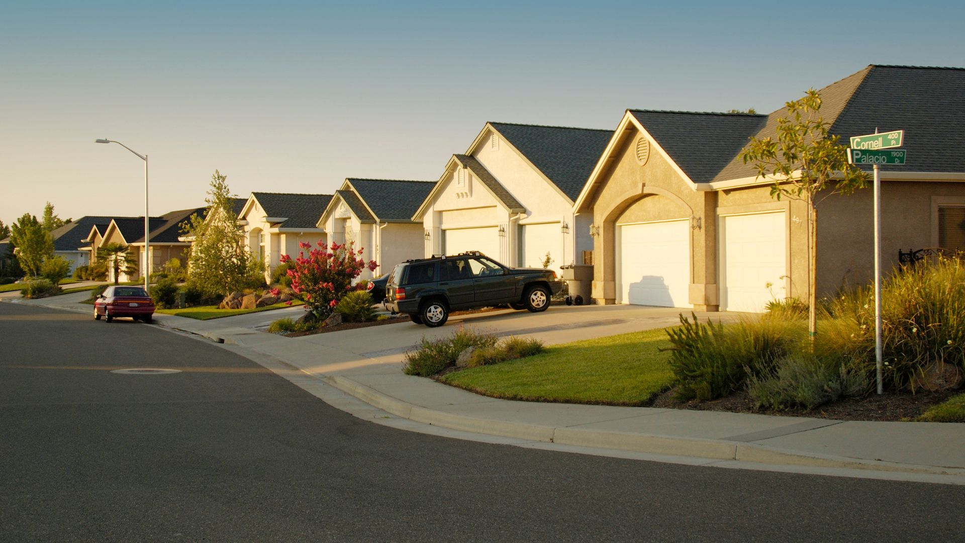 Suburban street with beige houses, green lawns, and parked cars at sunset