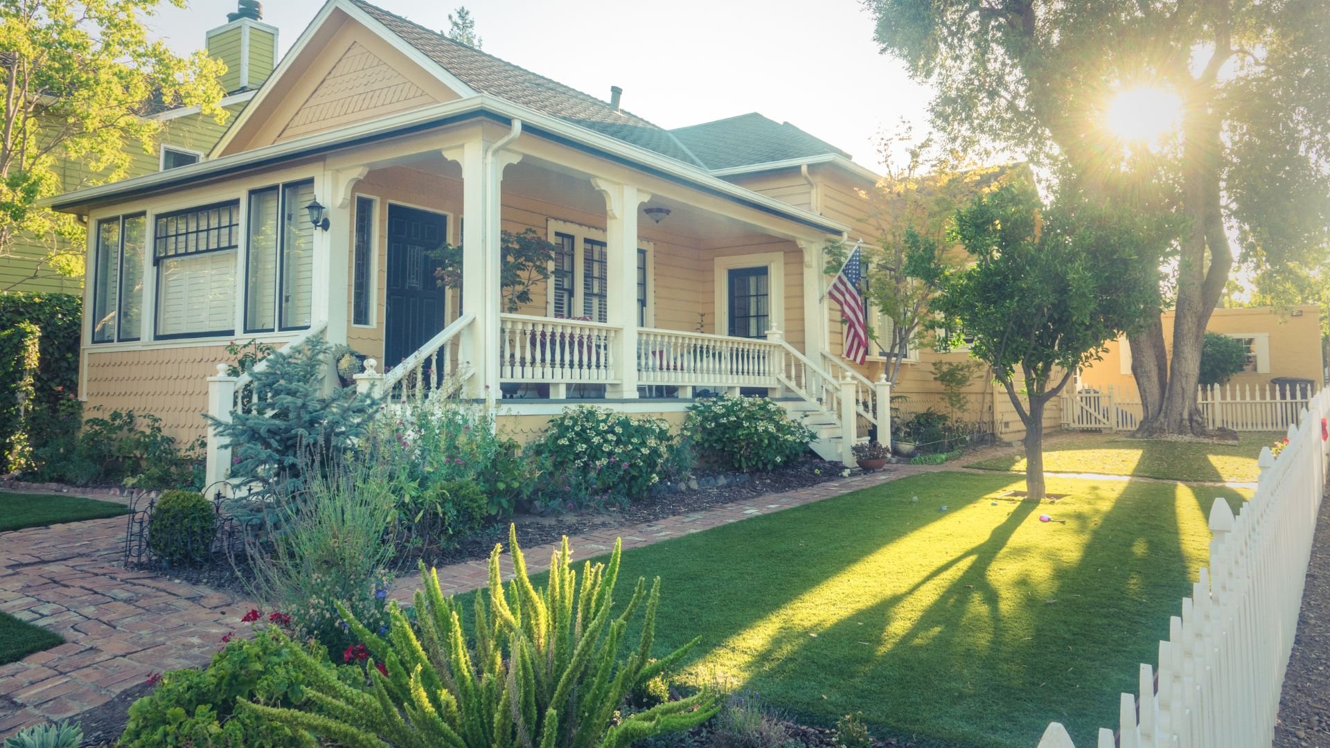Charming yellow cottage with white porch and American flag, sunlit garden